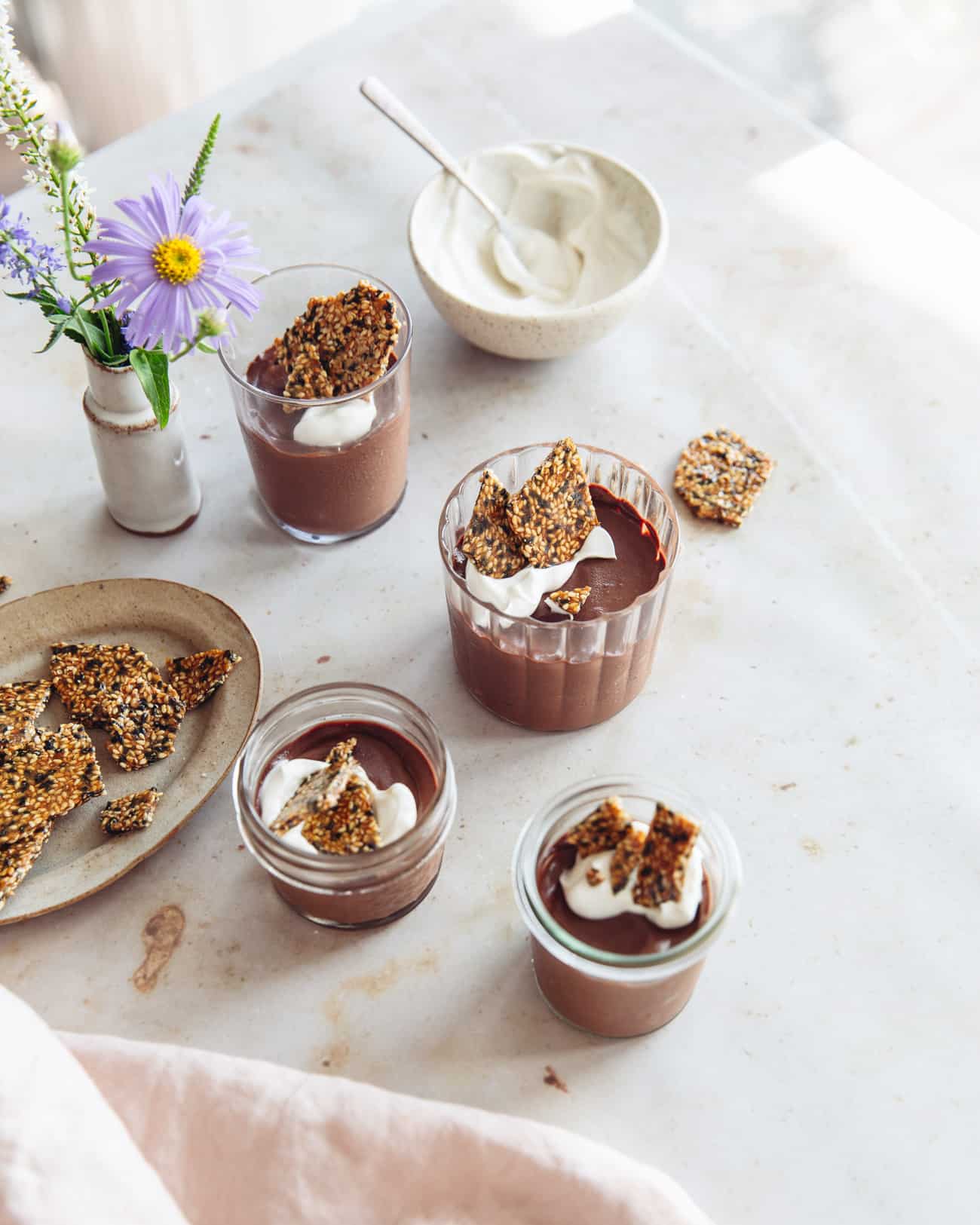 a cluster of glasses of chocolate tahini mousse with a bowl of creme friache, sesame brittle and a vase of flowers