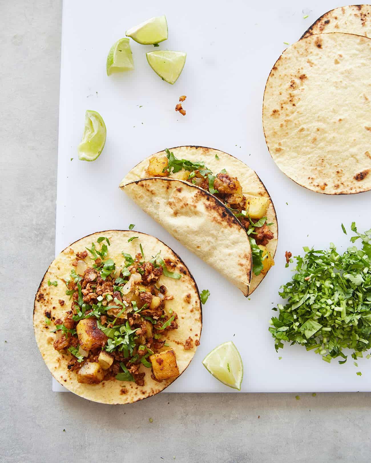 two vegan chorizo potato tacos on a chopping board with corn tortillas, cilantro and limes