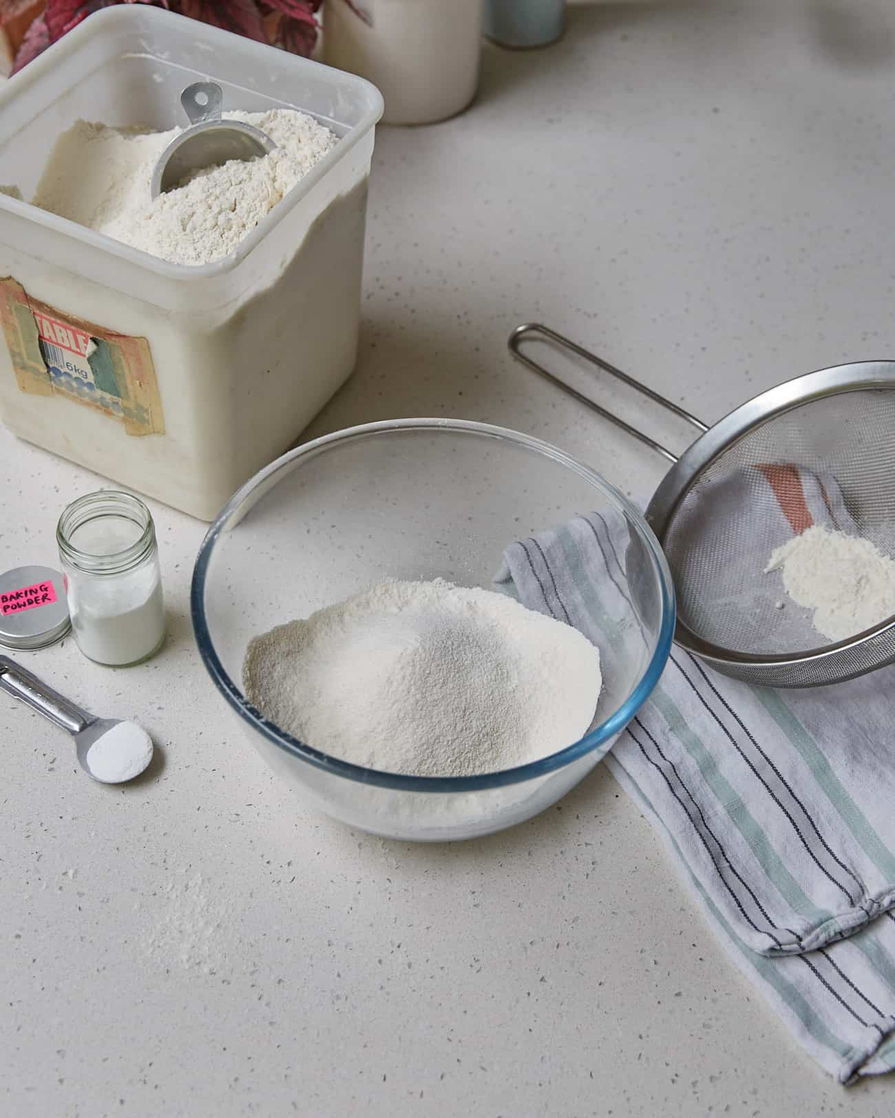 A bowl with homemade self-raising flour substitute, a sieve, some baking powder in a jar and a tub of plain flour